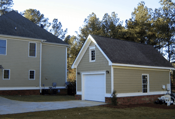 One car detached garage with tan siding and a brick base, next to the main house